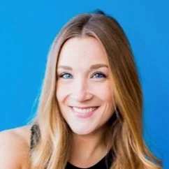A woman with long blonde hair and blue eyes smiles at the camera against a bright blue audiovisual backdrop, wearing a sleeveless black top.