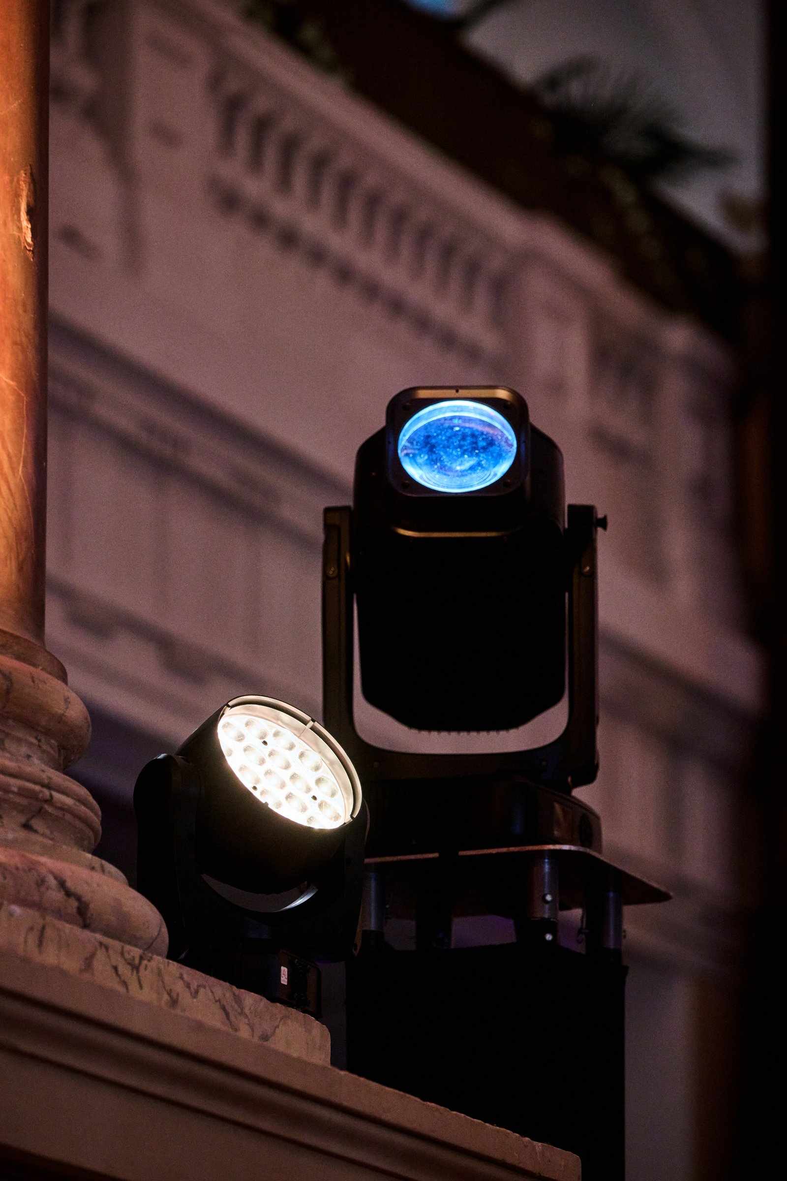 Two stage lights, one emitting blue and the other white, are mounted on a stand indoors against an ornate wall with decorative architectural details, creating an elegant setting ideal for various services.