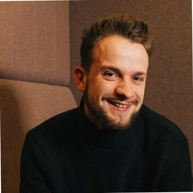 A man with short brown hair and a beard, wearing a black turtleneck sweater, smiles while sitting in front of a brown, padded audiovisual background.