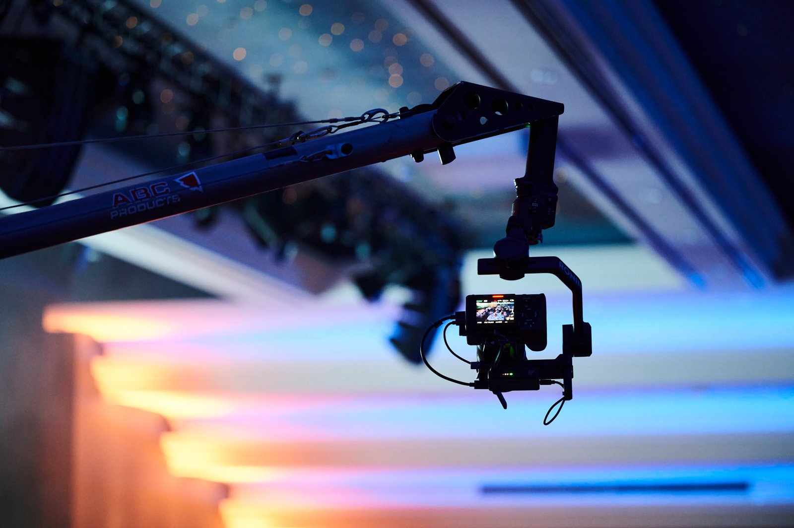 A camera mounted on a crane arm is silhouetted against a background of blurred, colorful stage lights in shades of blue, orange, and purple—highlighting the dynamic services behind live event production.
