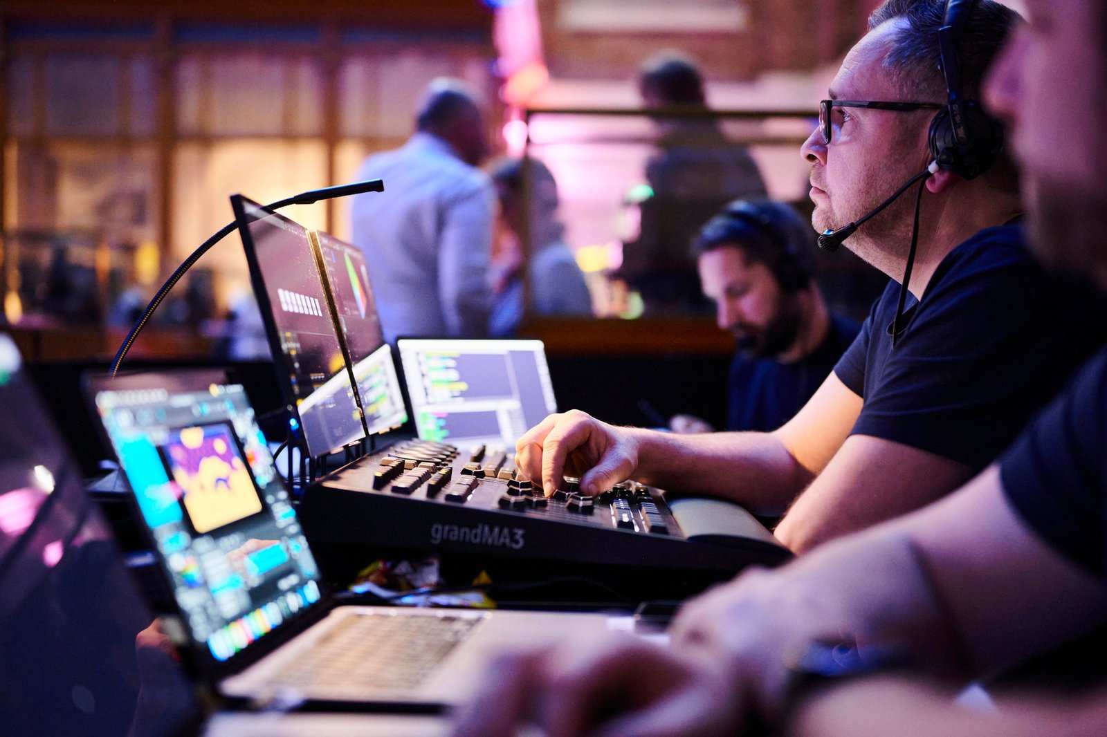 A man wearing a headset operates an audiovisual lighting control panel surrounded by laptops and monitors; other technicians work nearby in a dimly lit, busy event control room.