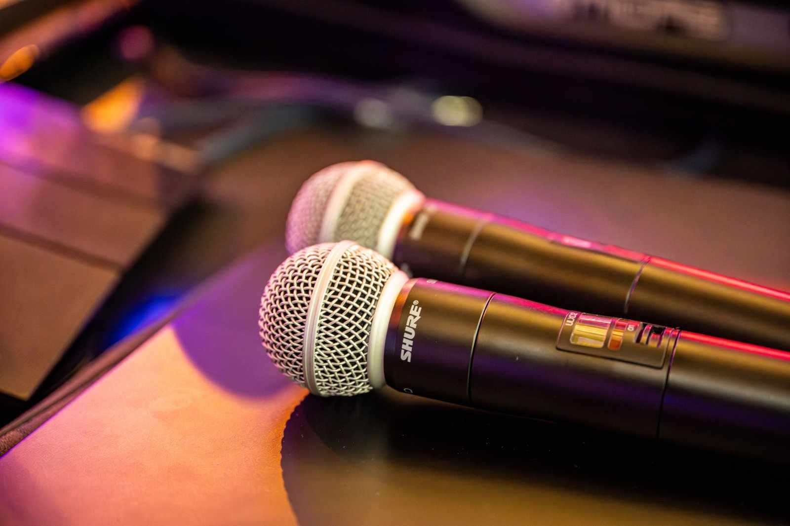 Two black wireless microphones with silver mesh heads lie side by side on a reflective surface, illuminated by soft, colorful ambient lighting—perfect for showcasing audio services in any vibrant setting.