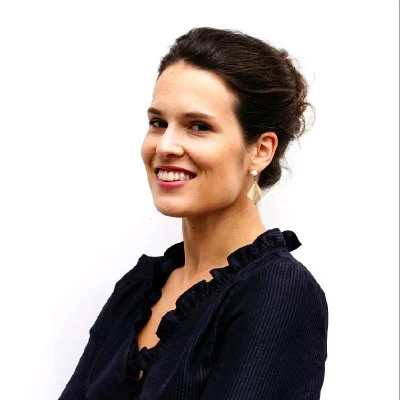 A woman with dark hair tied back, wearing a navy blouse with ruffled edges and gold earrings, smiles while posing for an audiovisual shoot against a plain white background.
