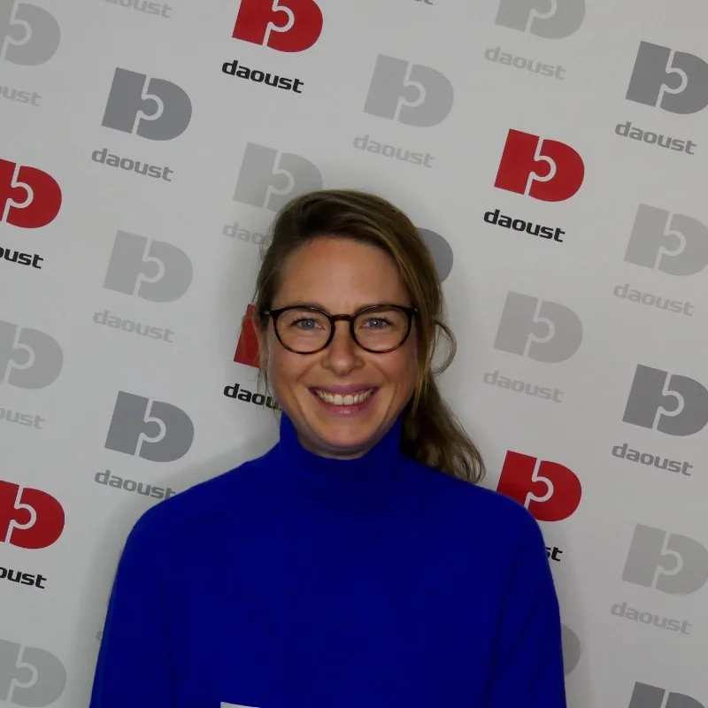 A smiling woman with glasses and a blue turtleneck stands in front of a backdrop featuring repeated “daoust” logos in grey, black, and red, at an audiovisual event.