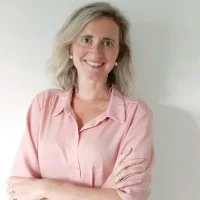 A woman with shoulder-length blonde hair and glasses, wearing a light pink blouse, stands against a plain white wall with her arms crossed and smiles at the camera, exuding confidence in an audiovisual setting.