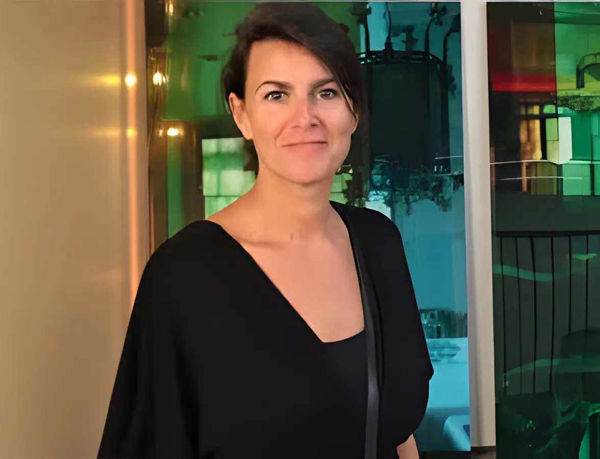 A woman with short dark hair and a black outfit stands indoors, smiling slightly. The background features colorful, reflective glass panels and soft, ambient lighting that evokes an audiovisual experience.