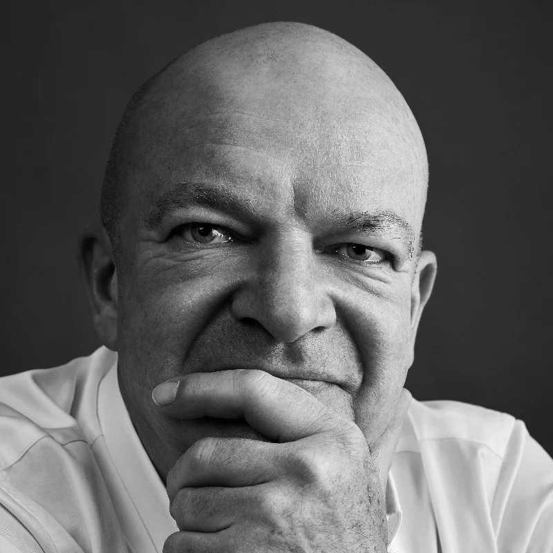 A bald man in a white shirt poses thoughtfully, resting his chin on his hand, gazing directly at the camera against a dark background, evoking a scene often found in sophisticated audiovisual productions.