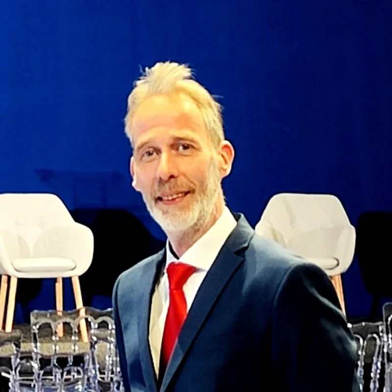 A man with short blond hair and a beard, wearing a navy blue suit, white shirt, and red tie, stands smiling in front of white chairs and an audiovisual setup against a blue background.