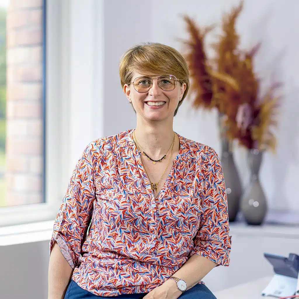 A smiling person with short light brown hair and glasses sits indoors by a window, wearing a patterned blouse and jeans. Decorative vases with dried plants are visible in the background, adding warmth to the scene about comfort and style.