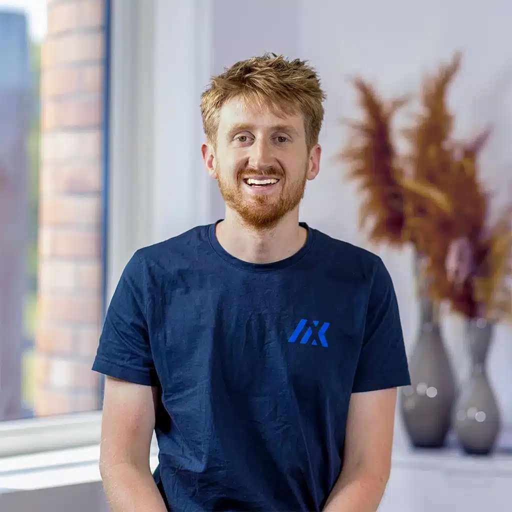 A smiling man with short, light brown hair and a beard sits indoors near a window with vases of dried flowers in the background, wearing a navy blue t-shirt with a blue logo—just about as relaxed as he looks.
