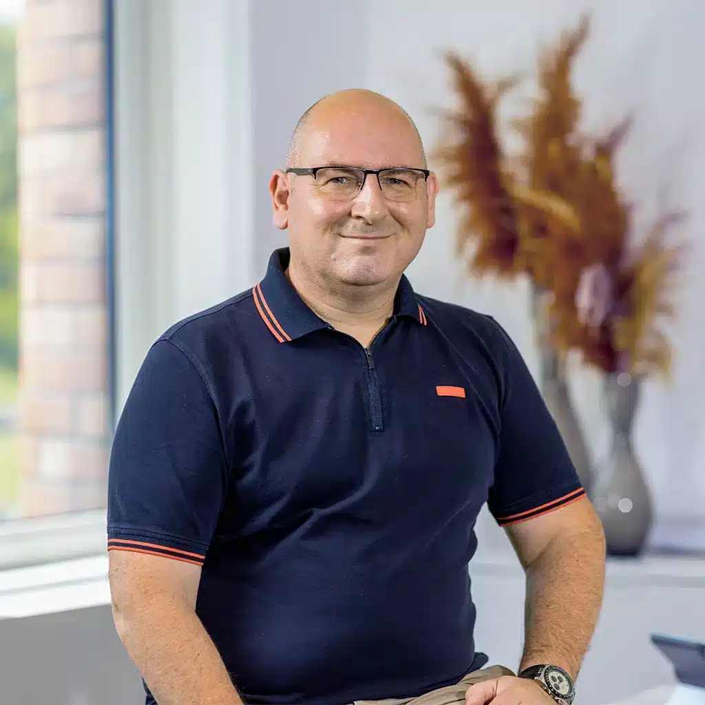 A man with glasses, wearing a navy polo shirt, sits and smiles indoors near a window. There is a vase with dried plants on a white table in the background, adding warmth to the scene about peaceful moments at home.