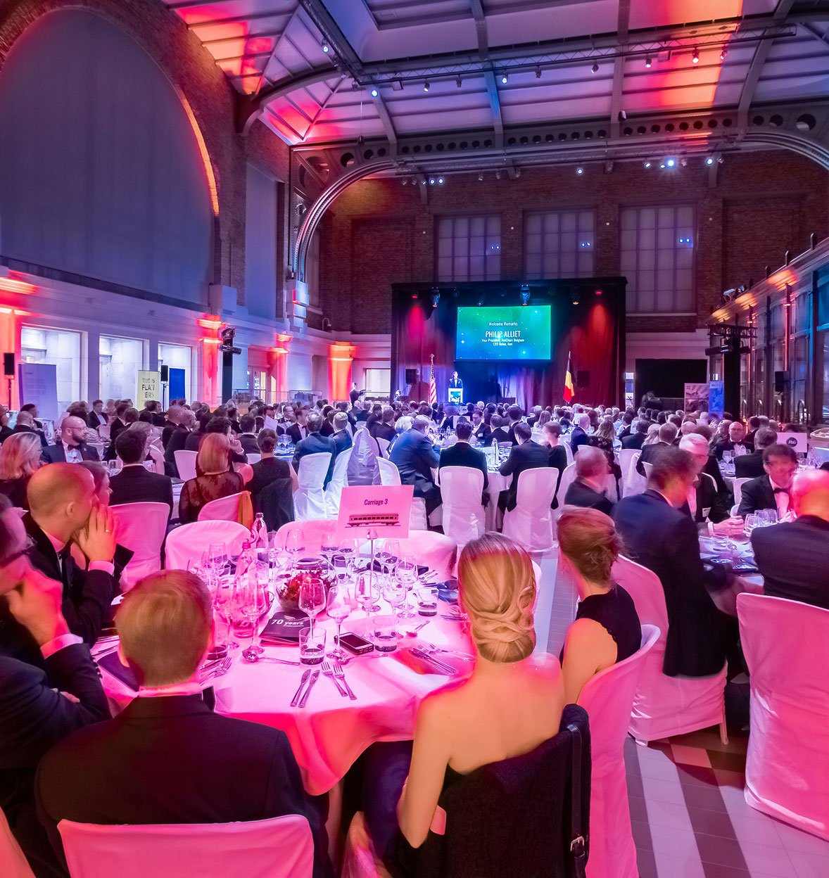 A large, elegant banquet hall filled with people seated at round tables covered in white cloths, attending one of the most anticipated events as they watch a speaker and screen on stage under colorful lighting. The atmosphere is formal and festive.