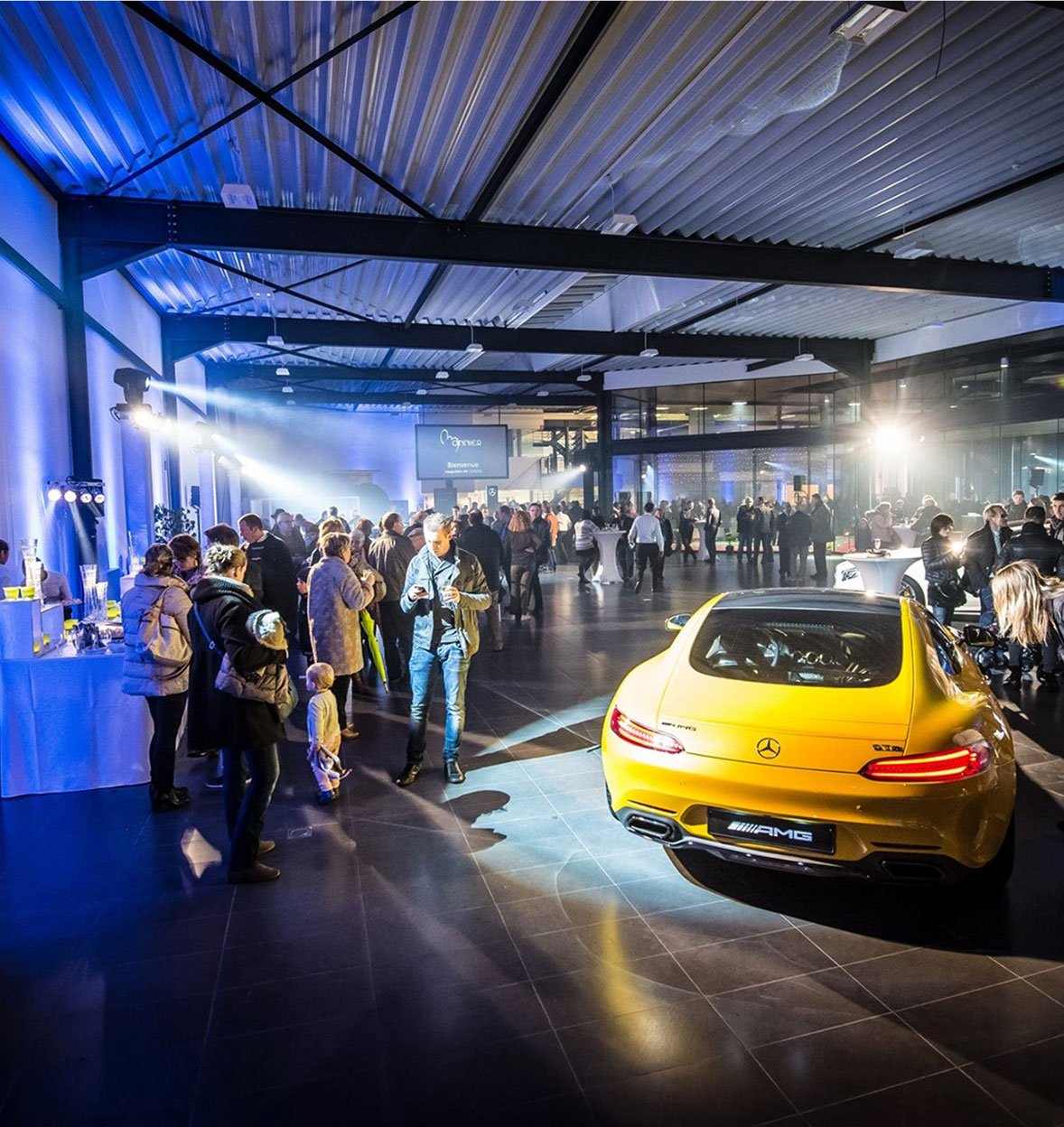 A crowd attends an indoor events showcase in a modern showroom, with a yellow Mercedes-AMG sports car displayed under bright lights in the foreground.