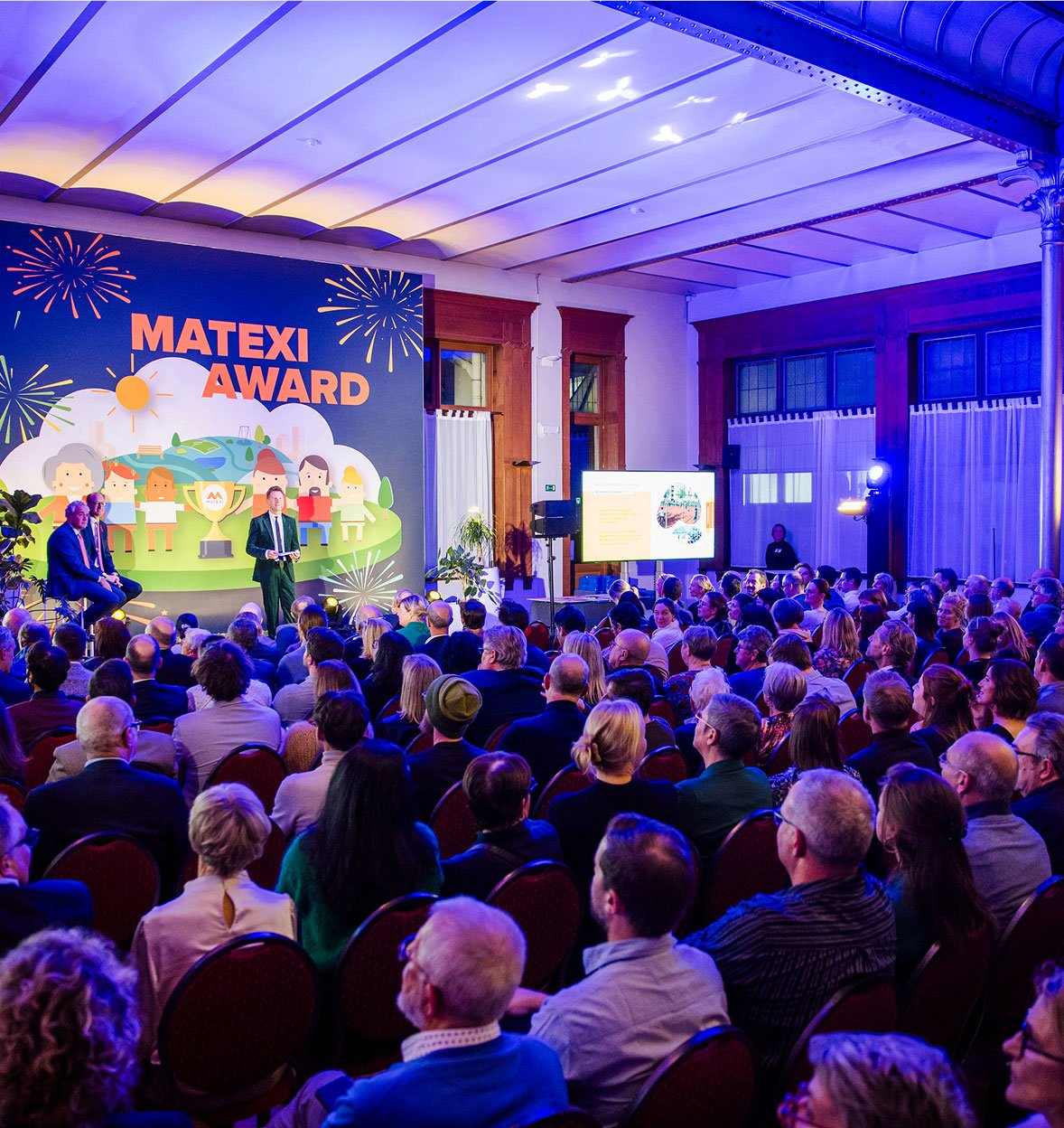 A large audience sits facing a stage at the Matexi Award events, with two speakers and a colorful backdrop featuring illustrated people, trophies, and fireworks. A presentation is being projected on a screen to the side.
