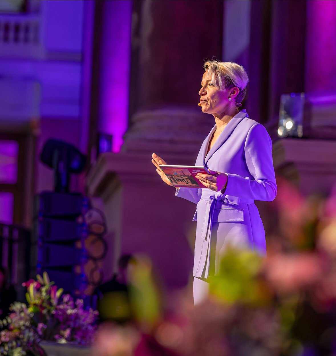 A woman in a light purple suit stands on stage at an event, holding a booklet and speaking under purple lighting, with blurred flowers in the foreground and ornate architecture in the background.