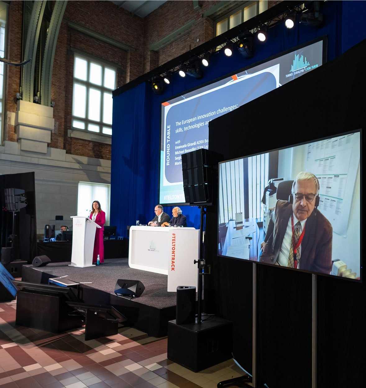 A woman speaks at a podium on stage beside two seated panelists during an events session. A screen displays a man in an office participating remotely, with large windows, a blue curtain, and a sign reading “INNO TRACK” in the background.