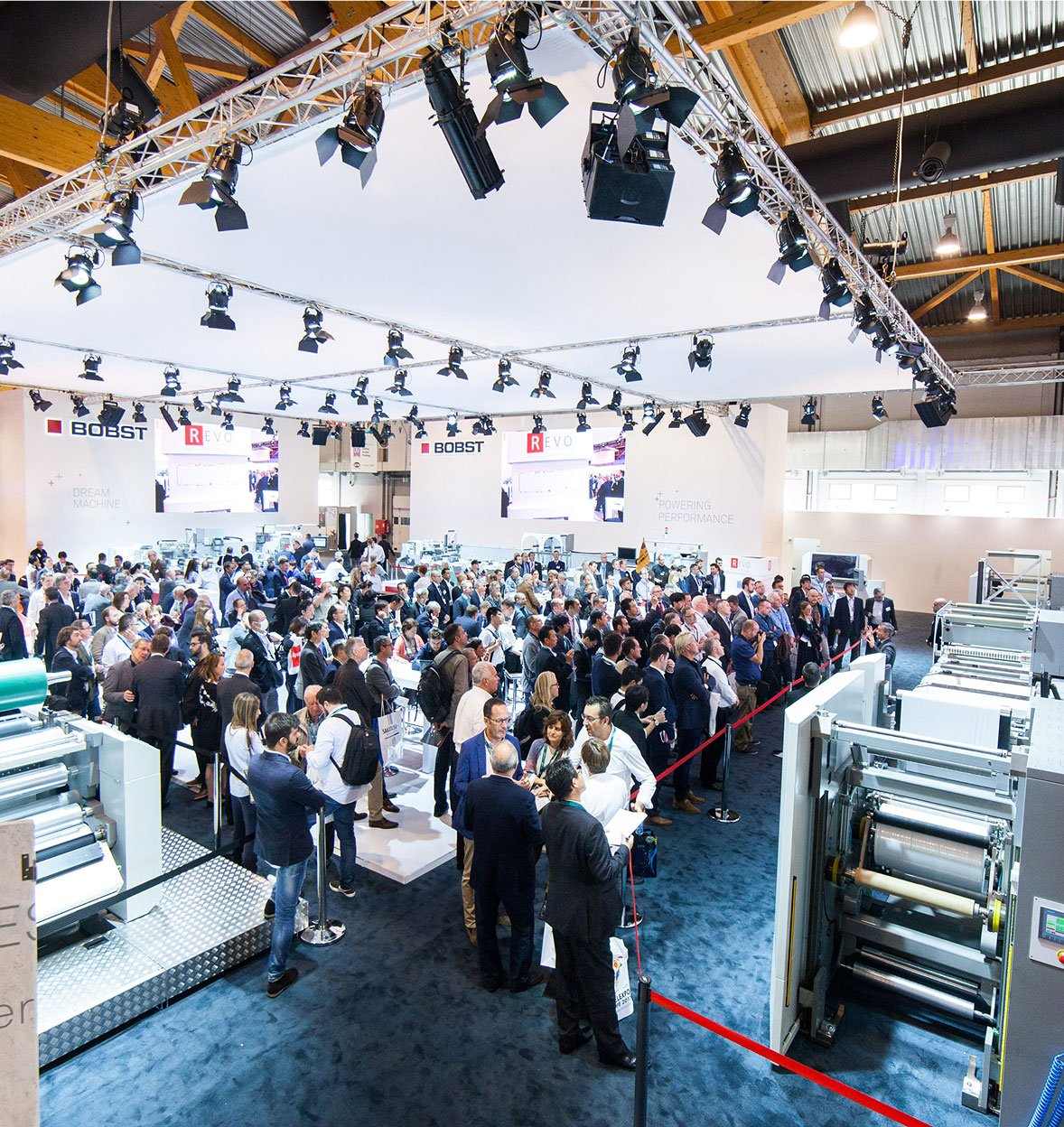 A large crowd of people attends a trade show or exhibition event, gathered around industrial printing or packaging machines, with bright overhead lighting and company booths in the background.