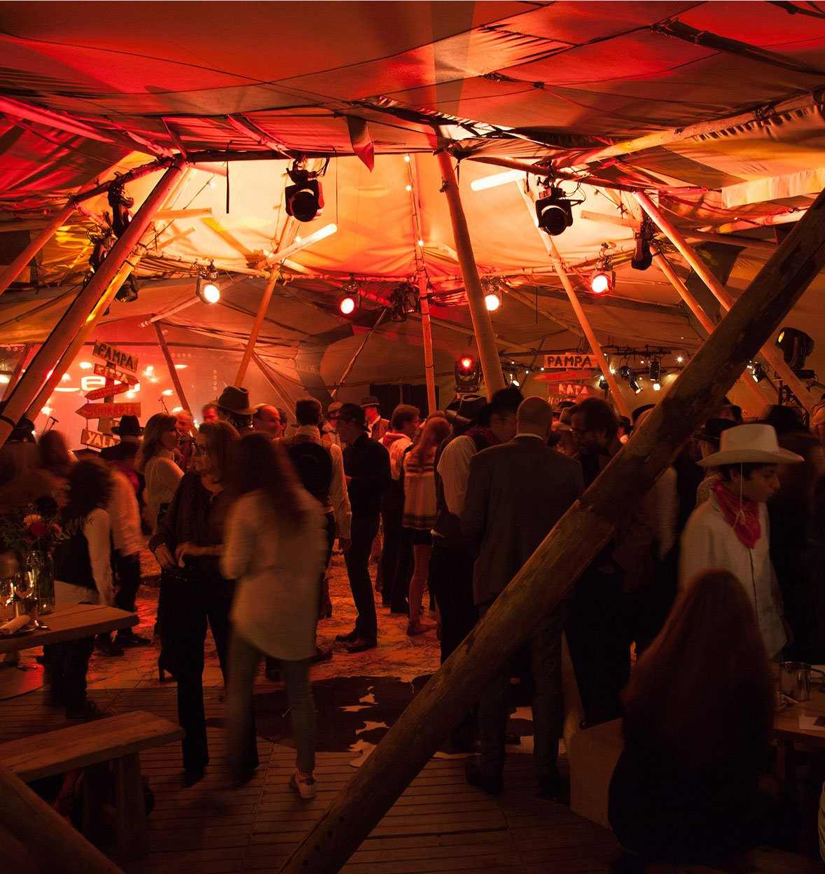 A lively indoor events party with people socializing under warm orange lights inside a tent-like structure with wooden beams, tables, and hanging lights, creating a festive and cozy atmosphere.