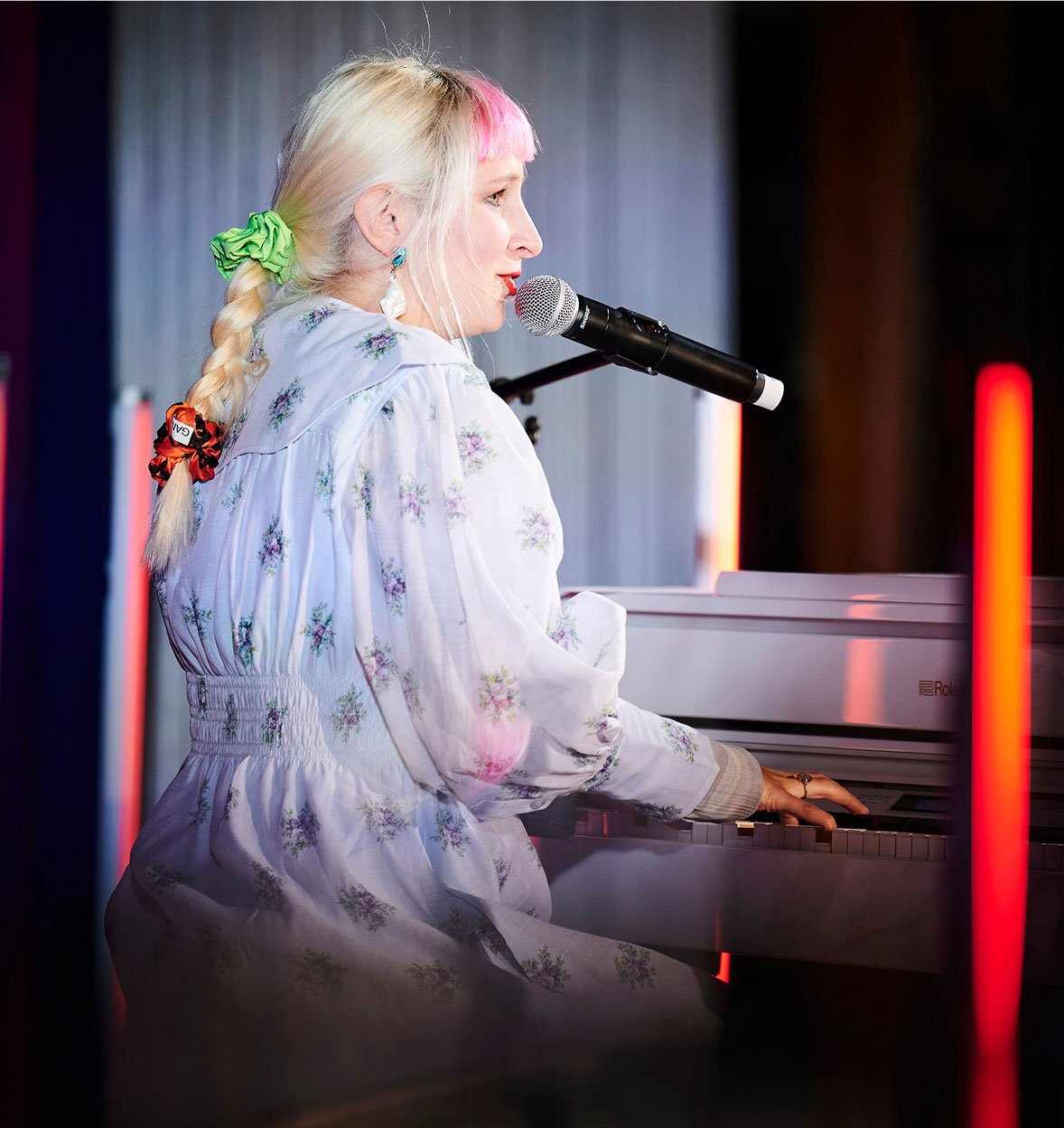 A person with long blonde hair, streaked with pink and tied in a braid, plays a white Roland piano and sings into a microphone. Dressed in a white floral dress, they perform at events amid colorful lights and soft illumination.