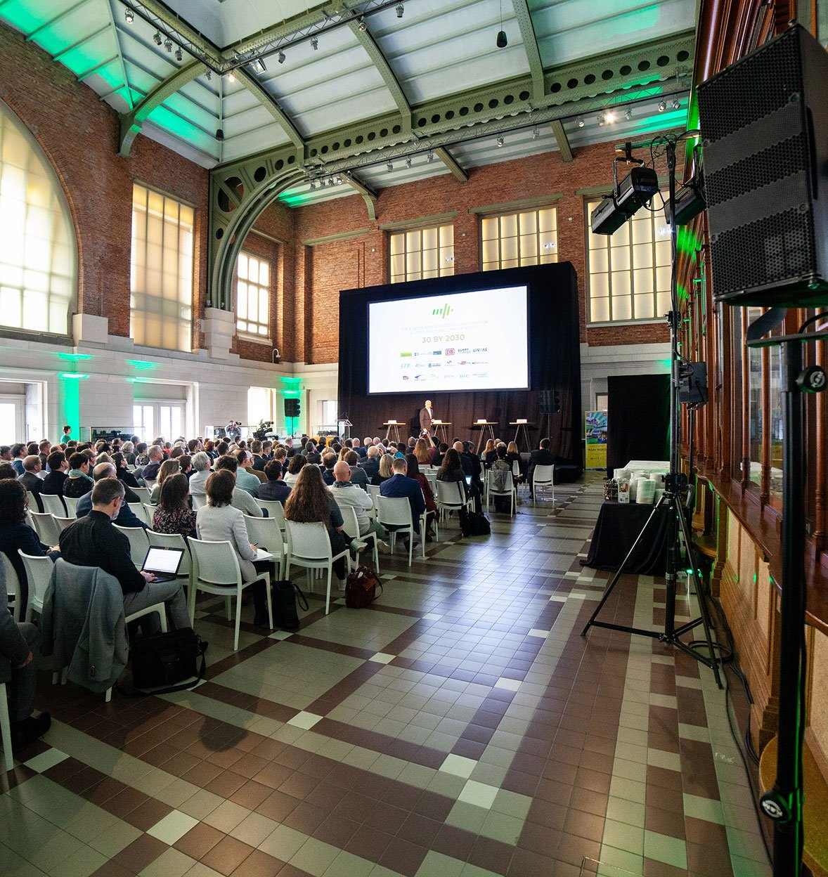 A large audience sits in rows facing a stage with a big screen in a spacious hall with high ceilings, brick walls, and arched windows—an ideal setting for events where a presenter stands at the front, delivering a talk or presentation.
